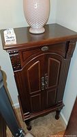 Wooden jewelry armoire standing upright, showing carved decorative detailing, cabinet doors with handles, and flat top surface with lamp on it.