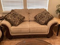 Photo showing beige loveseat with carved wooden base and two patterned throw pillows, positioned in front of window with blinds.