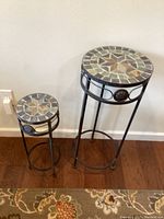 Photo of two round mosaic stone and wrought iron plant stands of different heights on a hardwood floor near a wall outlet.
