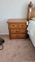 Wooden nightstand beside bed, showing two small upper drawers and two larger lower drawers with handles.