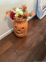 Front view of ceramic pumpkin jack-o'-lantern vase with fall flowers, showing smiling face and floral arrangement.