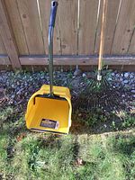 Photo of large yellow plastic dustpan with long metal handle and black grip standing on grass next to metal leaf rake with wooden handle.