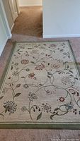 Full view of the rectangular floral pattern area rug laid on a carpeted floor showing the green border and flower motifs.
