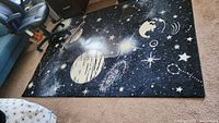 Top-down view of the full throw rug placed on carpeted floor, showing space-themed pattern with planets and stars.