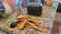 Photo of black toaster and a pile of wooden spoons, spatulas, a black plastic slotted spoon, and a metal grater on a granite countertop.