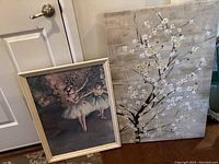 Both framed art pieces leaning against a wall near a door: one with ballerina print in a frame, the other textured tree art on blocked vinyl-like material.