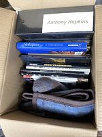 Top-down view inside box showing black Anthony Hopkins binder, multiple books about films and Shakespeare, and a folded cloth item at bottom