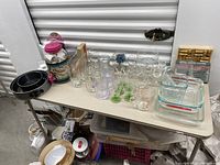 Wide view of Pyrex baking dishes, glassware including wine glasses, and baking pans on plastic folding table.