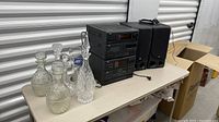 Photo showing five glass decanters, two beer steins, a black compact stereo system with multiple components and two matching black speakers on a table