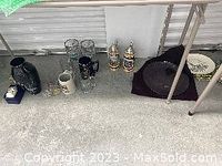 Photo showing black vase, two German lidded beer steins, clear glassware, crystal platter, and Coca Cola platter under a table.