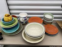 Photo showing a variety of Fiesta dinnerware items stacked on a table including plates, bowls, and a small pitcher, exhibiting assorted colors and patterns.