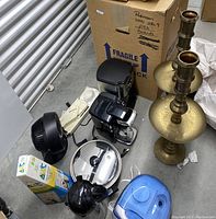Photo showing the entire lot of kitchen and household accessories on the floor of a storage unit, including two tall brass candle holders, two coffee makers (Cuisinart and Keurig), electric fry pan with lid, small fan, humidifier, hand slicer, over-the-door traction unit, and carton of paper cups.