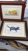 Two framed watercolor paintings, one of a brown animal curled in grass, the other of a black cat, placed on floor with surrounding items.