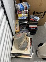 Photo showing a pile of about 35 LP records in sleeves with visible wear and several stacks of DVDs including titles such as 'Shallow Hal' and other movie collections, positioned near a cardboard box.