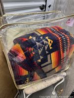Two wool blankets in clear protective case. One is a solid cream color, the other with red, black, yellow geometric pattern design.