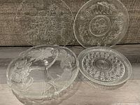 Four clear glass trays displayed on floor, featuring different floral and etched designs, various sizes