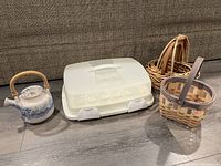 Overview of all items in lot on floor: ceramic tea pot, white plastic cupcake carrier, and three woven baskets.