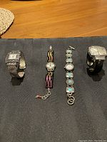 Four ladies watches laid out on dark fabric showing different styles including silver band, beaded chain, turquoise stones, and wide black band with rectangular face.