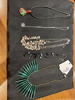 Photo showing 5 necklaces laid on black fabric: handblown glass pendant on black cord, silver chain with Tree of Life pendant, multi-chain necklace with silver and copper beads, black bead necklace, and green pendant necklace.
