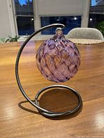 Full view of the purple glass orb suspended on a silver curved metal stand on a wooden table.