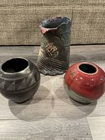 Photo showing all three pottery vases placed on the floor against a beige couch: large multicolor Raku vase, red MCM vase, and black vase with geometric patterns.