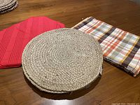 Photo shows a round jute placemat centered between one red solid fabric placemat and one fall plaid fabric placemat with autumn colors on a wooden surface.