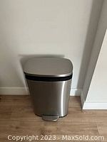 Image of a stainless steel rectangular trash can with foot pedal placed against a white wall on a wooden floor.