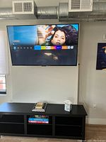 Wall-mounted 55-inch Amazon Fire TV Omni Series smart TV turned on and displaying the Alexa smart TV home screen interface. Room features a black TV console underneath with books and board games.