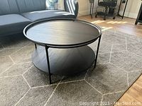 Photo showing round coffee table with raised tray edge, two tiers in dark oak finish with matte black metal frame in a living room setting.
