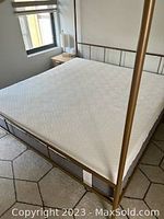 White textured king size mattress on a metal four-poster bed frame in a bright room with a window and a nightstand with lamp.