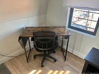 Office chair positioned at a wooden desk beside a window, showing black mesh back, armrests, adjustable height base with rolling wheels.