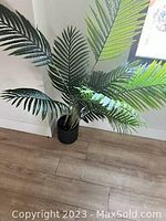 Photo of artificial green palm-like plant with multiple leaves in a simple black pot placed on a wooden floor near a wall and window.