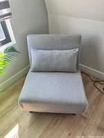 Light gray tri-fold convertible sofa bed chair with removable pillow shown folded as a chair on wooden floor in room corner.