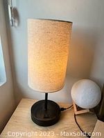 Photo of a cream fabric shade table lamp with a black base placed on a wooden surface, lamp is lit showing warm light.