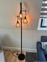 Tall black industrial floor lamp with three lit bulbs in protective cage wire shades, placed beside a sofa.