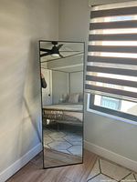 Full length mirror leaning against a wall in a room with wood flooring and black window blinds.