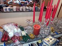 Overview of various candles and glass candle holders on counter including red taper candles, green taper candles, tea lights, a large red jar candle, and clear glass holders.
