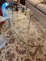 Oval glass coffee table with gold metal frame and curved legs shown in living room setting.