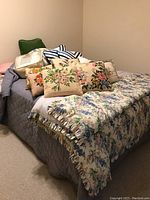 Wide view of bed showing multiple colorful and floral throw pillows, a down blanket in plastic packaging, and a floral comforter with a ruffle edge.