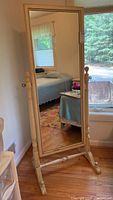 Full front view of a full-length floor mirror standing on a wooden floor, showing its beige painted wooden frame and spindle style vertical supports.