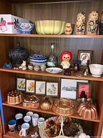 Shelf with Russian nesting dolls, ceramic containers, Raggedy Ann doll, framed art prints, coffee mugs, Bundt molds, and decorative basket.