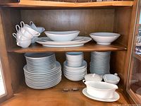 China set displayed in wooden cabinet showing stacks of plates, bowls, cups, saucers, and serving pieces