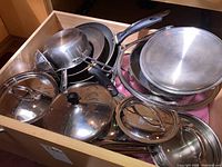 Drawer with assorted pots, pans, and lids of various sizes and types, showing surface wear and use marks.