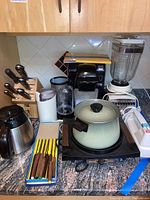 Overview of kitchen countertop showing all items in the lot including Keurig coffee maker, knife block, Black & Decker coffee pot, fondue maker, blender, and cutlery.