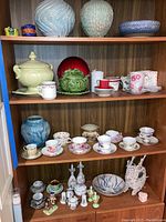 Wide shot of shelf with various ceramic pots, bowls, tea cups, salt and pepper shakers including large blue pots, green cabbage pot, red dish, and multiple floral tea cups on saucers