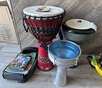 Full set photo showing large red djembe drum, smaller aluminum djembe, bongo drums, and instruction CDs on floor.