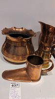 Photo showing the large scalloped copper bowl, two copper jugs, and the lance-shaped ale warmer with handle and base, placed closely together on a plain surface.