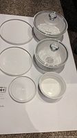Three ceramic CorningWare casserole bowls; two have clear glass lids, and three white plastic covers are laid out.