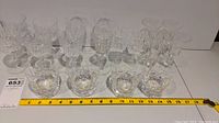 Full view of 15 crystal glasses including tumblers and stemware arranged in rows with a measuring tape showing length for scale.