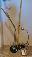 Photo showing two metal desk lamps side by side with measuring tape for height reference. One lamp has a black base and dual necks, the other has a silver base and a flexible neck.